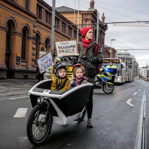 Das Foto stammt von einer FridaysForFuture Demo. Zu sehen ist eine Mutter mit zwei Kindern im Fahrrad-Anhänger, die Demoschilder hochhalten.